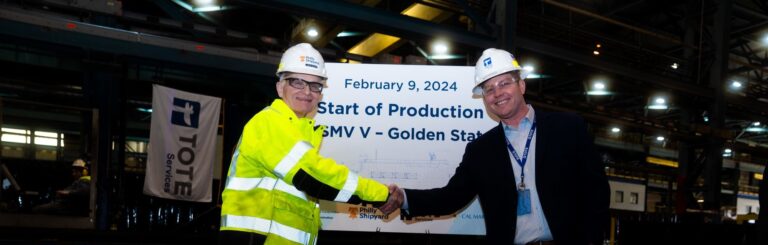 Two men in safety gear shaking hands at steel cutting ceremony for NSMV ship.