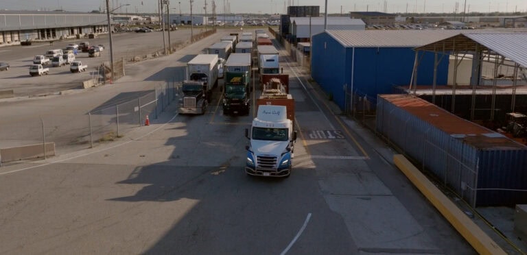 A busy truck yard with multiple trucks parked and moving near blue and red warehouse buildings.