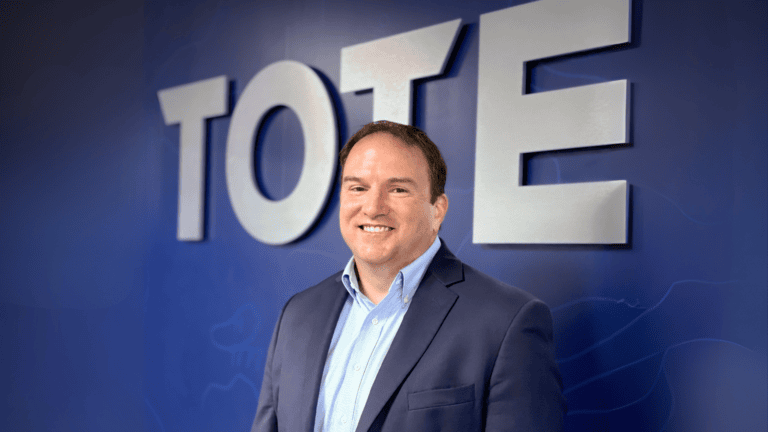 A smiling man in a suit standing in front of a blue background with the word "TOTE" in large letters.