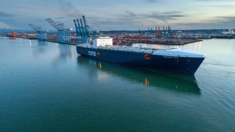 Large navy vessel named TOTE in a harbor with cranes in the background.
