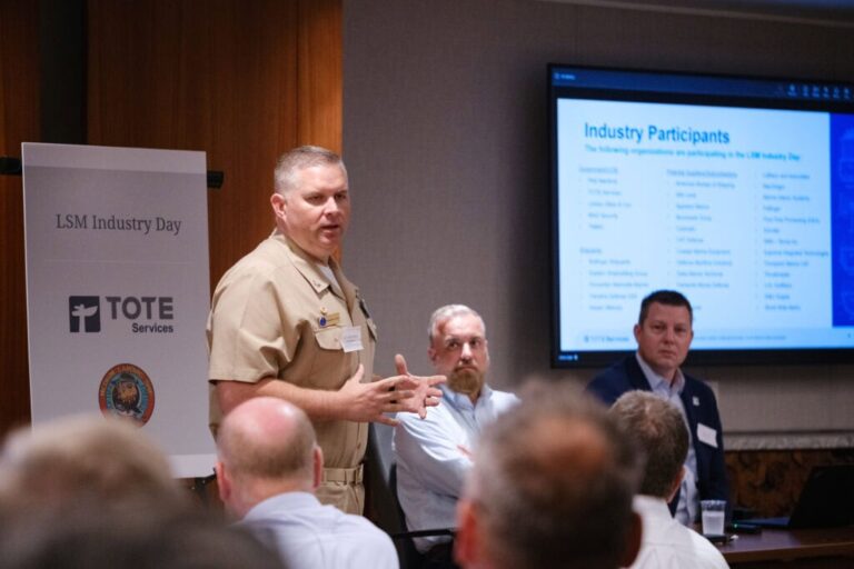 Man speaking at a conference with a presentation screen behind him.