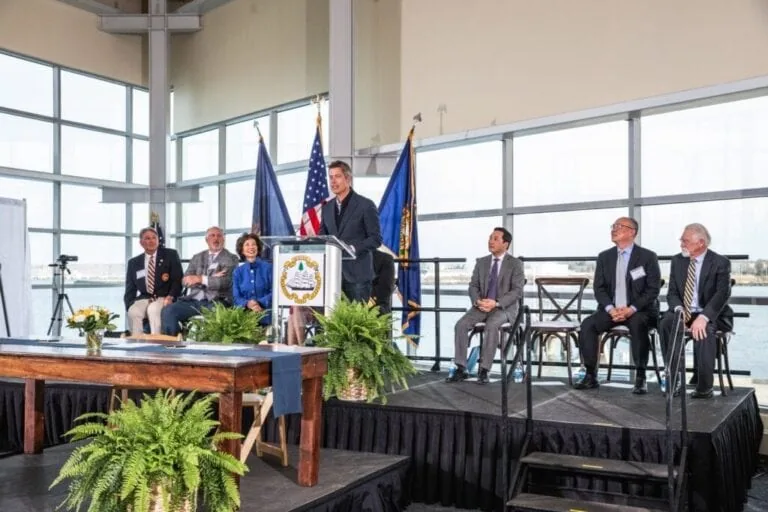 Man speaking at podium during formal event with seated officials behind him.
