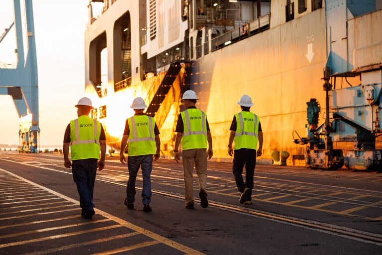 Workers inspecting a ship at a dock during sunset, wearing safety vests and helmets.