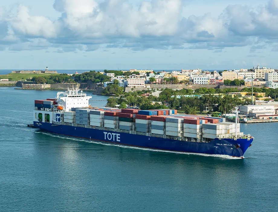 TOTE cargo ship sailing in a harbor with a cityscape in the background.