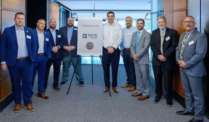 Group of nine men in business suits standing in a room with a sign that reads 'TOTE'.